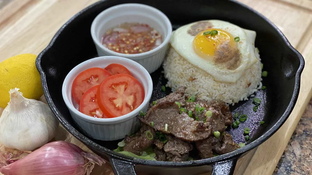 Beef Tapa prepared in an iron skillet with a side of rice with an egg over the top